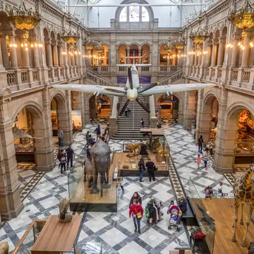 People walk below an airplane hanging in a large atrium with a black-and white floor in a museum in Glasgow, Scotland.