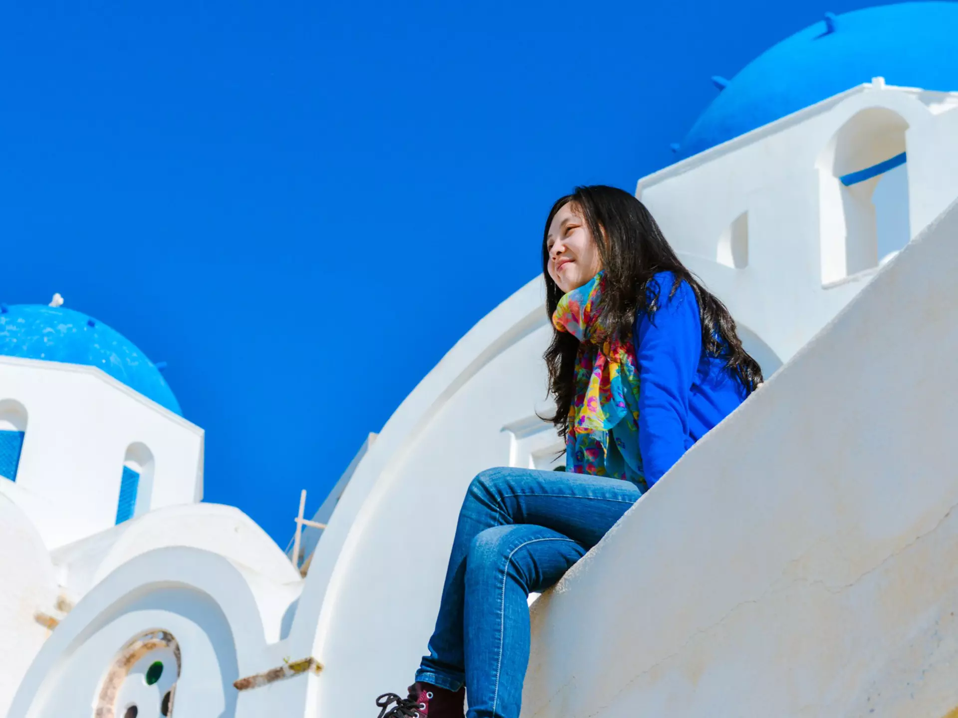 A woman sitting on the edge of a rooftop in Oia town and enjoying the view of Santorini.