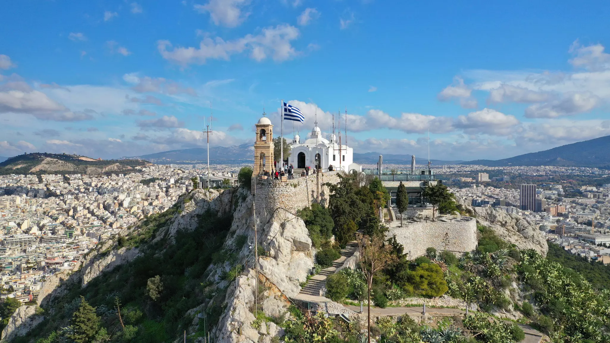 Aerial shot of chapel on top of a rocky hill with beautiful deep blue sky and clouds and a city stretching out into the distance.