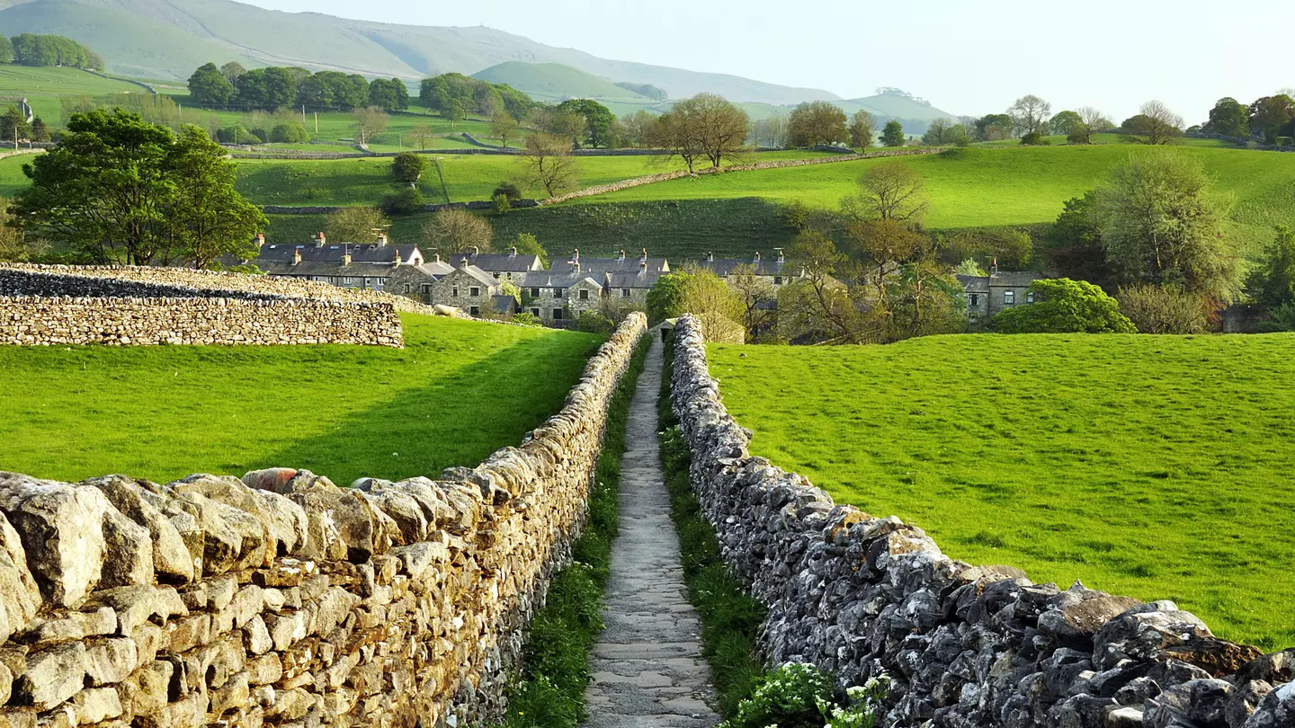 A path, lined with drystone walls, stretches out ahead through countryside leading to a village of stone houses.