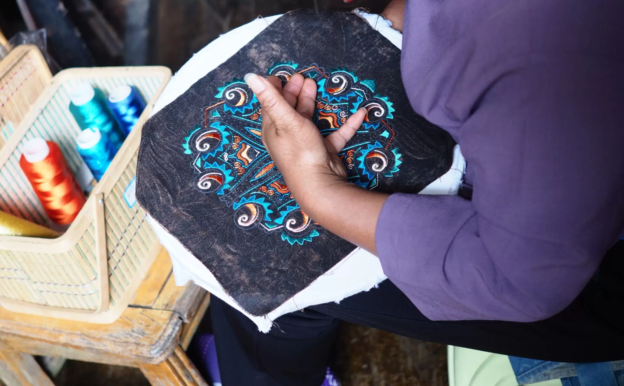 Overhead view of a woman's hands embroidering a colorful spiral motif onto dark fabric, beside a basket of teal and orange thread spools.
