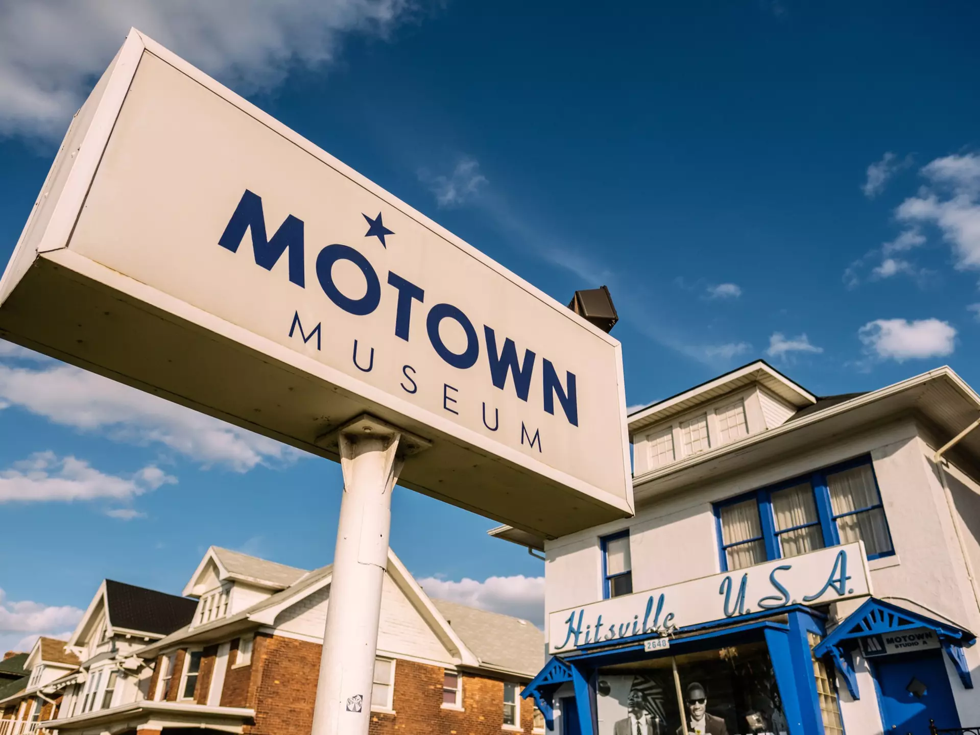 Exterior of a white building with blue trim and the words "Hitsville U.S.A" above the entry and a large sign in front reading "Motown Motown" on a sunny day.