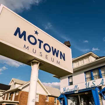 Exterior of a white building with blue trim and the words "Hitsville U.S.A" above the entry and a large sign in front reading "Motown Motown" on a sunny day.