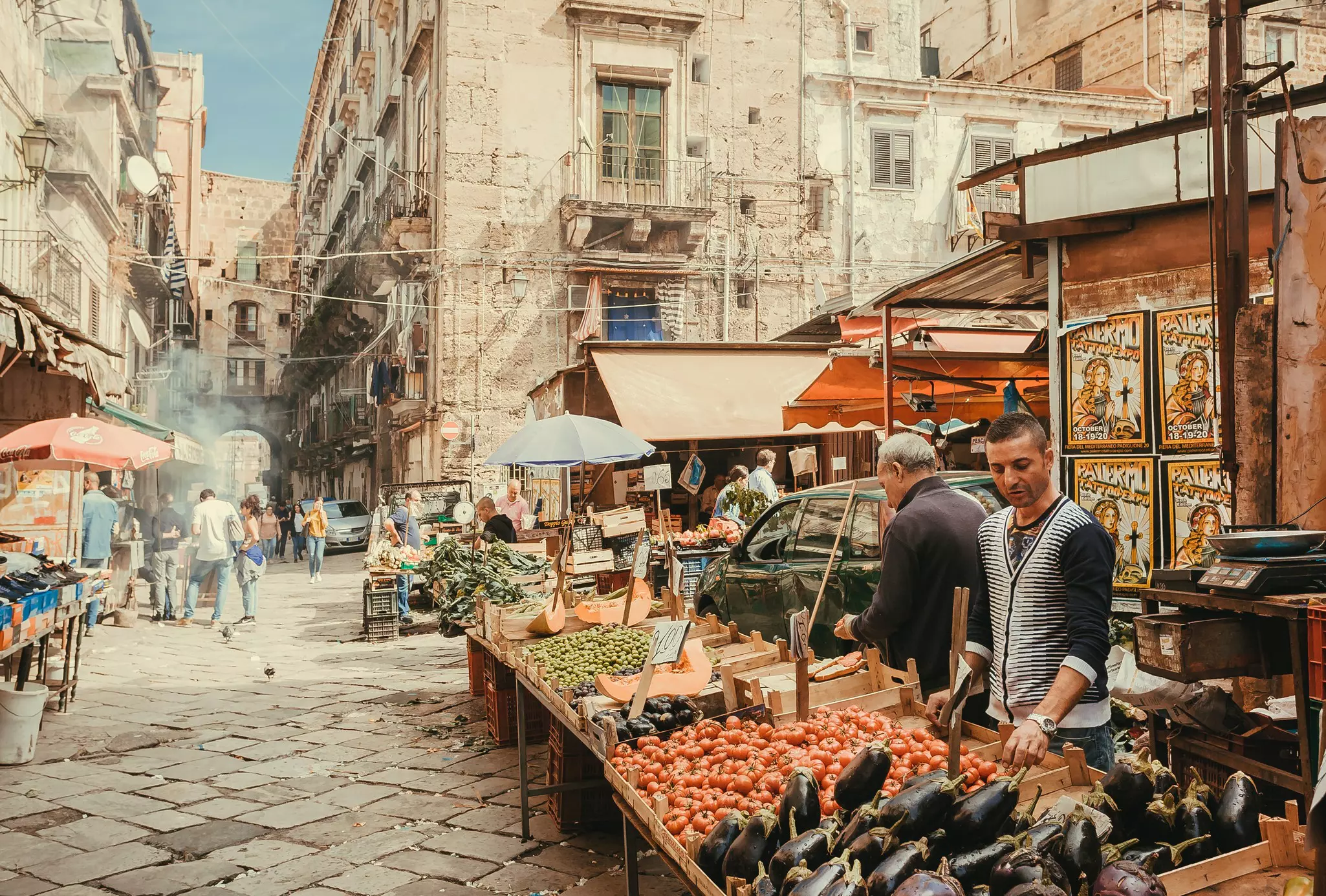 Two men sell tomatoes, eggplant and other fresh vegetables from an open stall in a narrow, pedestrianized, cobbled street in an old city.