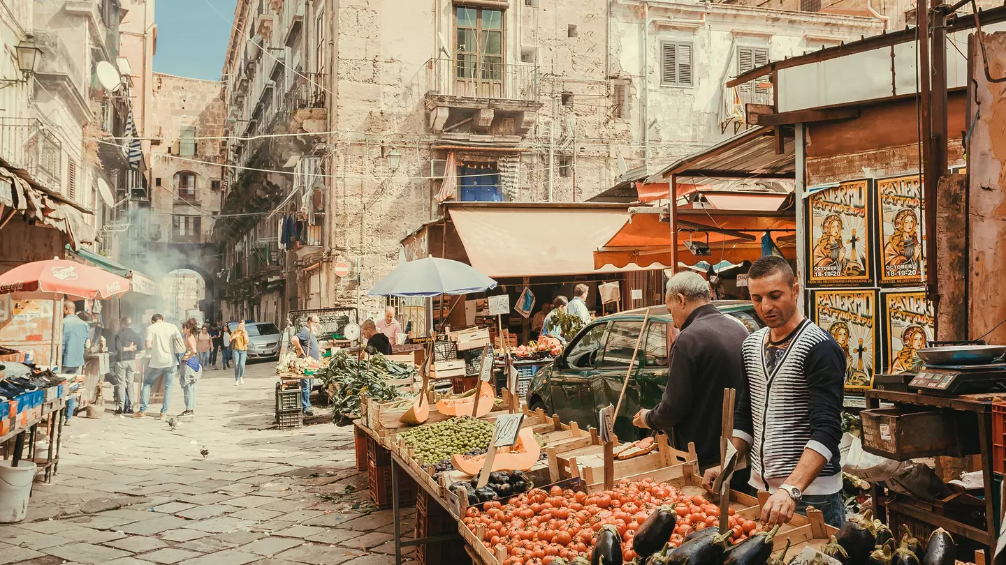 Two men sell tomatoes, eggplant and other fresh vegetables from an open stall in a narrow, pedestrianized, cobbled street in an old city.