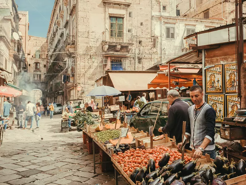 Two men sell tomatoes, eggplant and other fresh vegetables from an open stall in a narrow, pedestrianized, cobbled street in an old city.