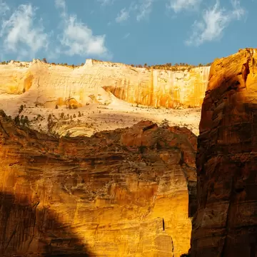 Sheer orange and cream sandstone cliffs glowing in low sunlight, half in shadow, beneath a blue sky with scattered cloud.