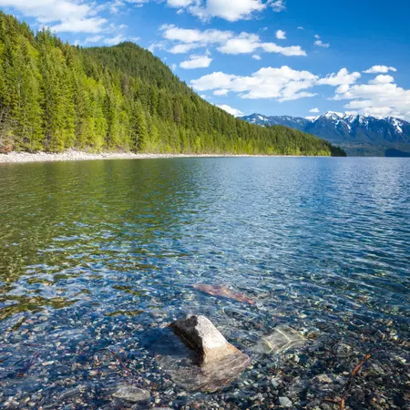 A lake with forested mountain to the left and snow-covered rocky peaks in the far distance on a sunny day.