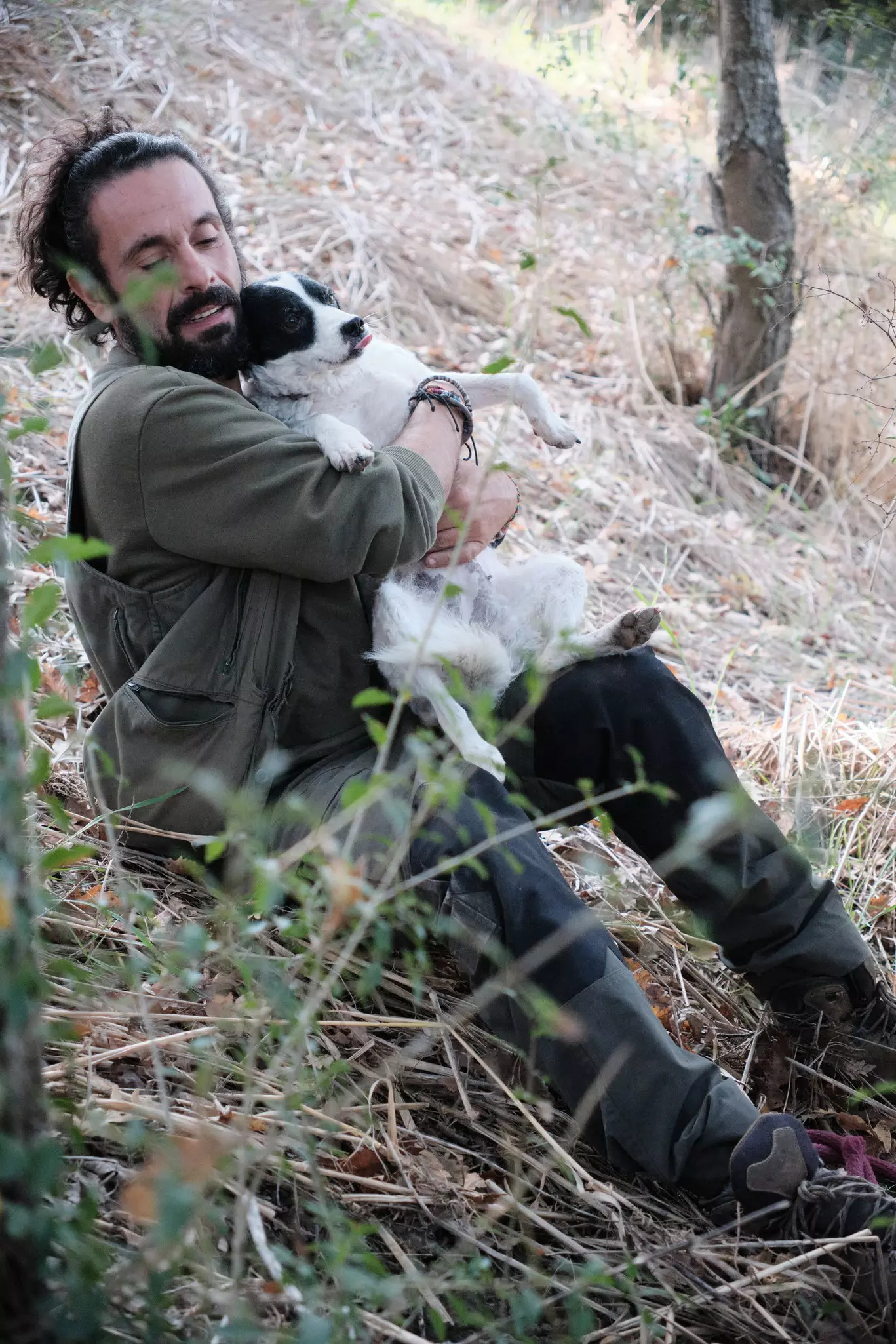 A man sits on the grass in woodland cuddling a wriggling black and white dog.