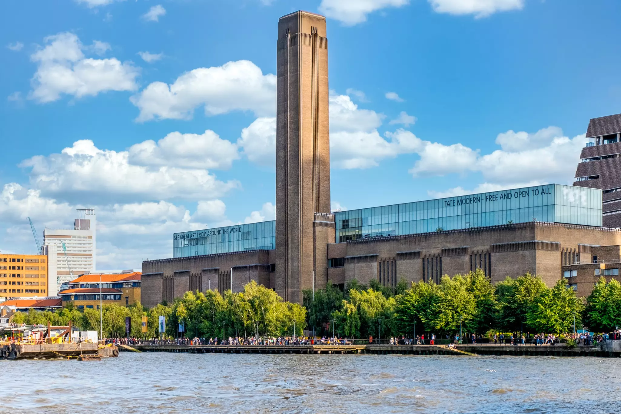 Wide shot of stone building with tall tower and glass roof reading "Tate Modern – Free and Open to All" on a partly sunny day.