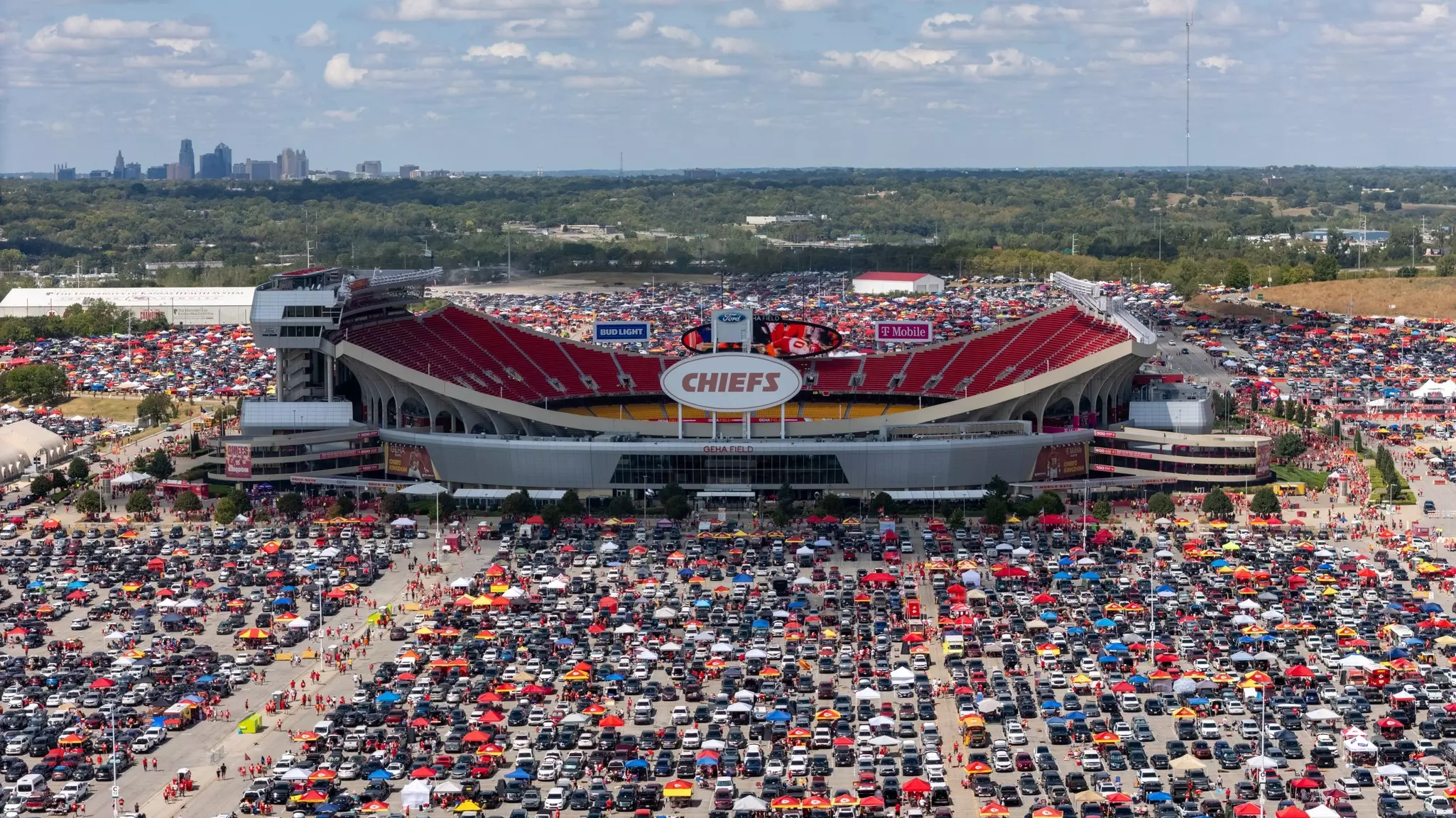 Aerial view of GEHA Field at Arrowhead Stadium in Kansas City with carpark full of cars and city skyline in the background