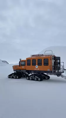 A woman stands with her arms out wide in front of a sign that reads "Union Glacier Antarctica"