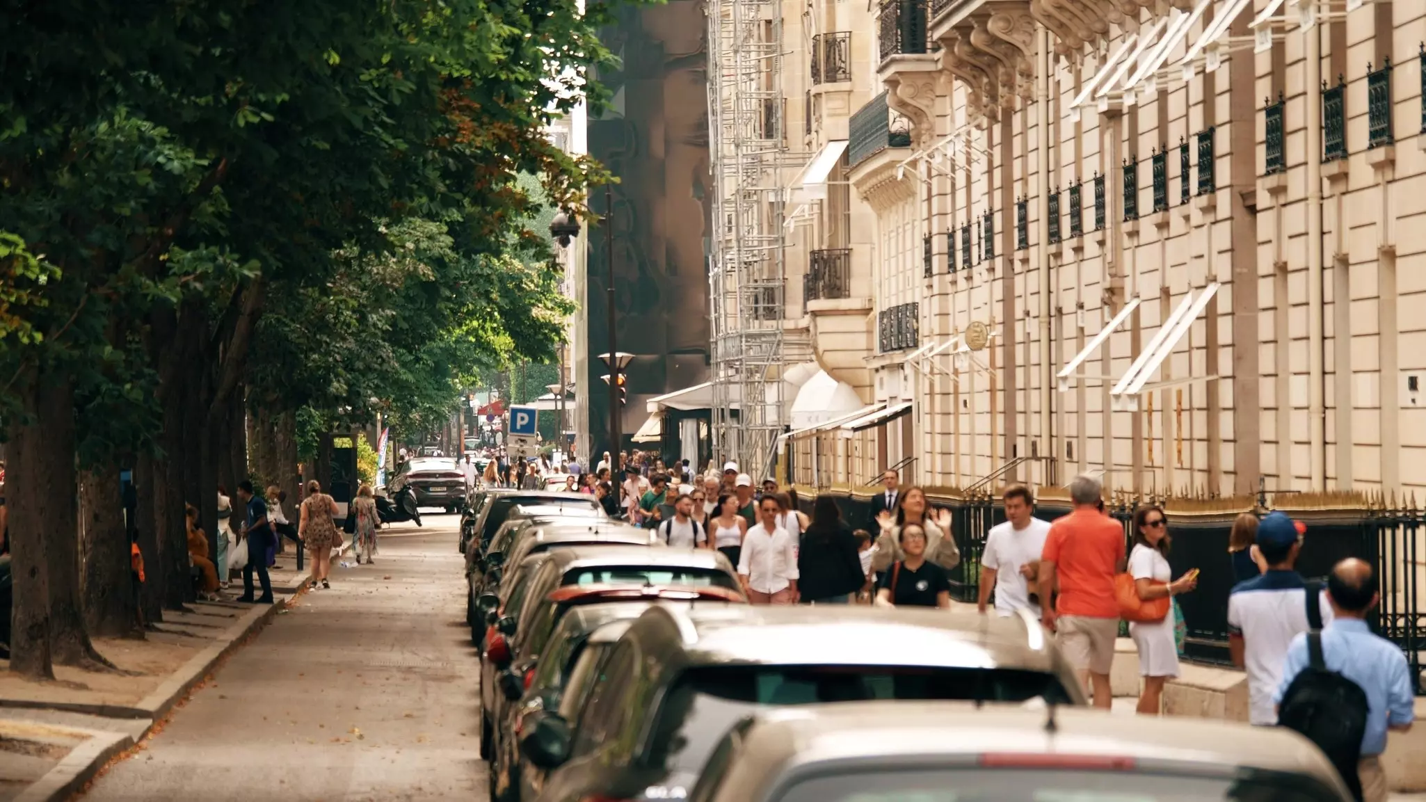 People walk on a sidewalk along a street in Paris with luxury shops on one side and trees on the other.