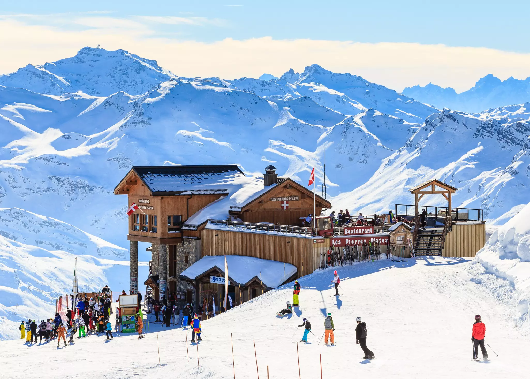 A wide view of skiers at the top of a mountain. A hut with a restaurant and terrace overlooks a valley, with snowy peaks all around.