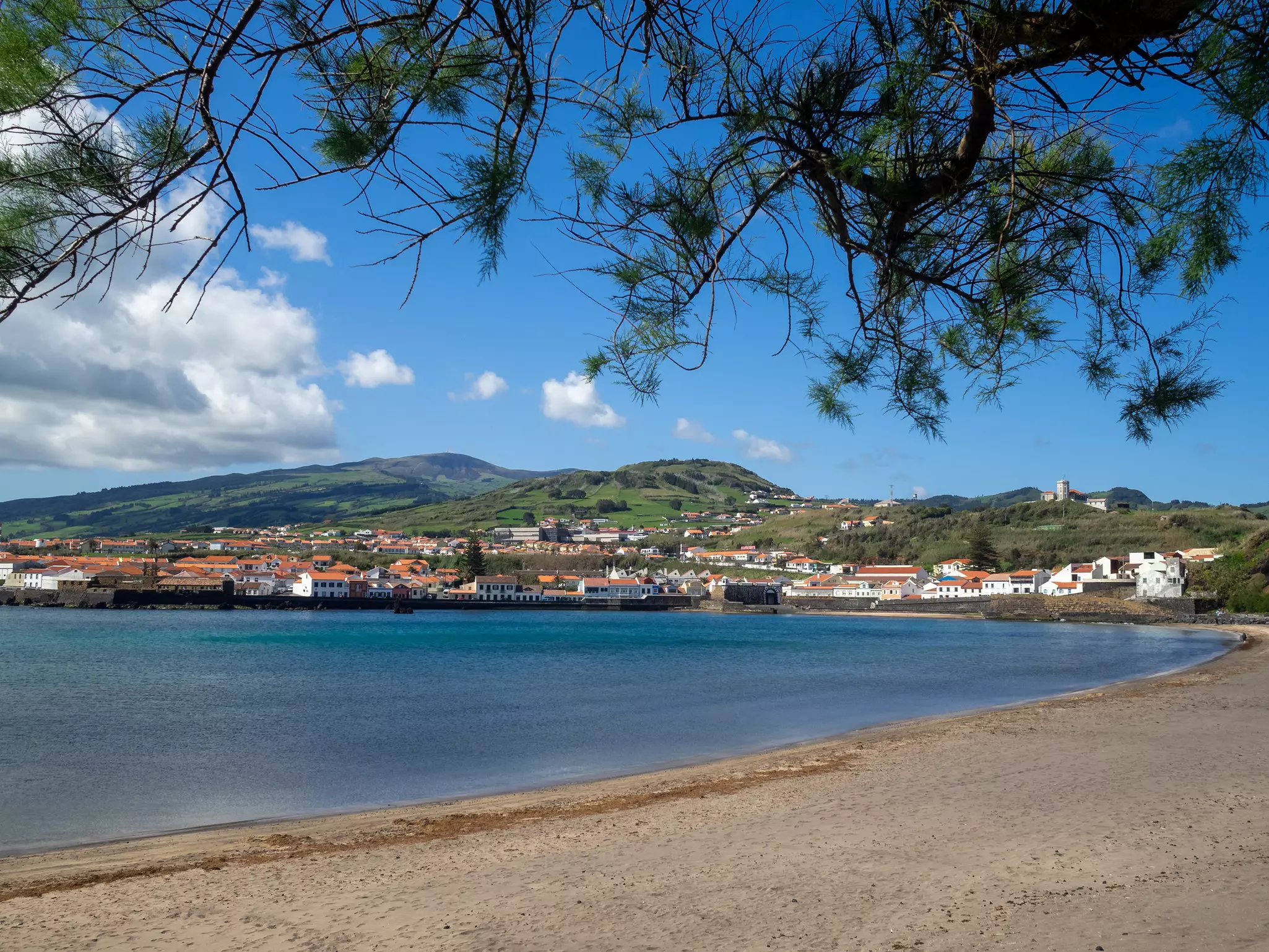 Curved sandy beach with ocean to the left and small town and green hills in the distance on a mostly sunny day.