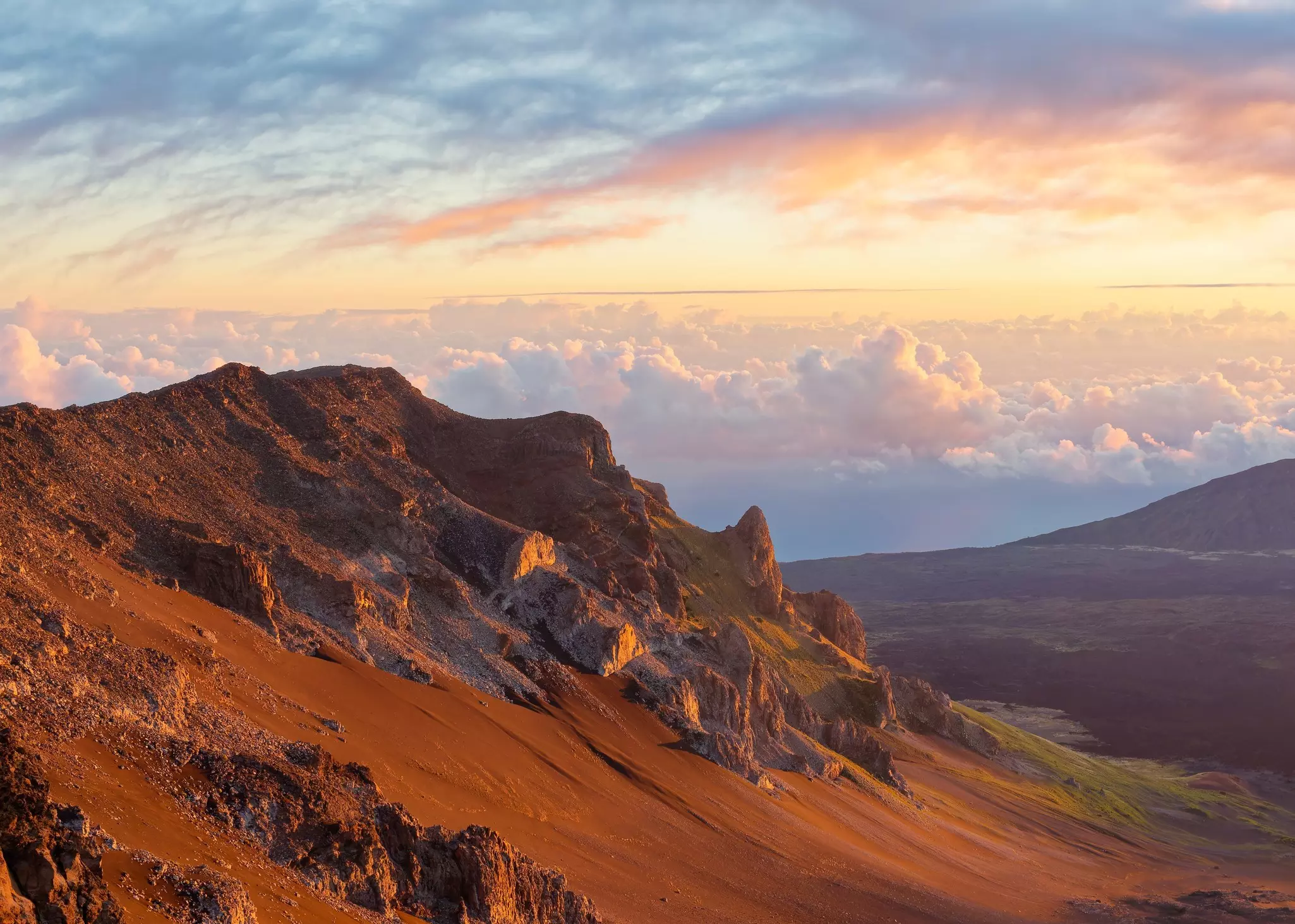 Sunrise along jagged mountains surrounded by red earth and clouds in the distance.