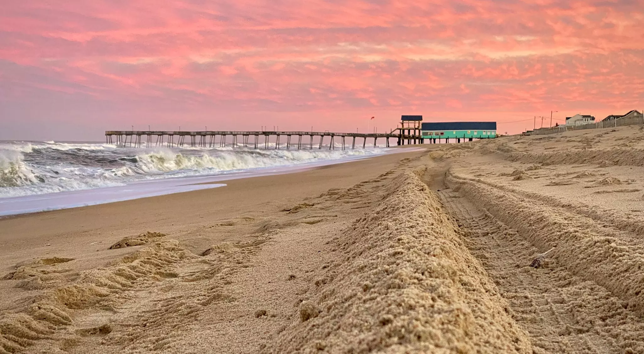 Sandy beach with coral-colored sunset sky and a pier in the background.