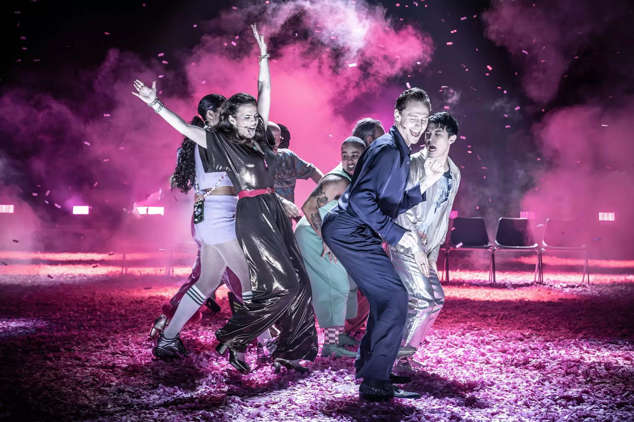 A group of dancers perform together on a stage covered in pink confetti and lit with pink light