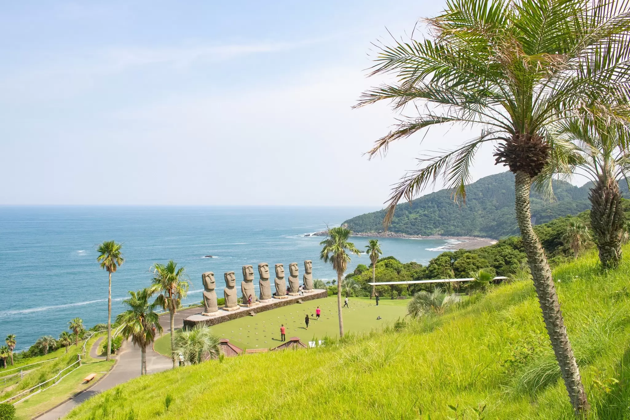 People clustered in front of seven monumental figure sculptures set on green grass; the ocean is beyond.