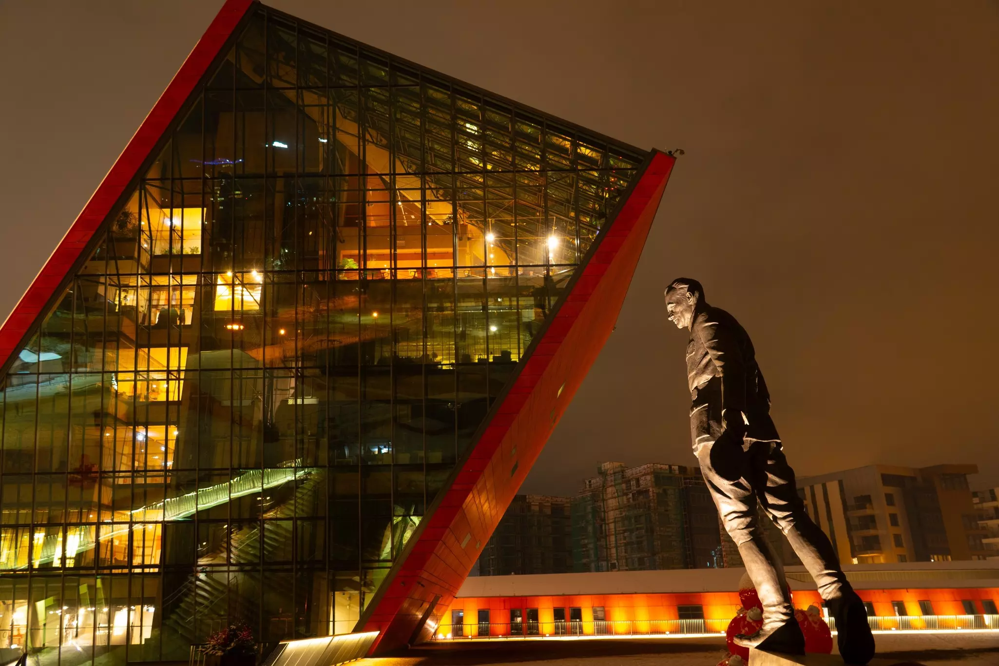 A nighttime view of a modern museum whose exterior is made of glass and red panels. A statue that leans forward is seen in the foreground.