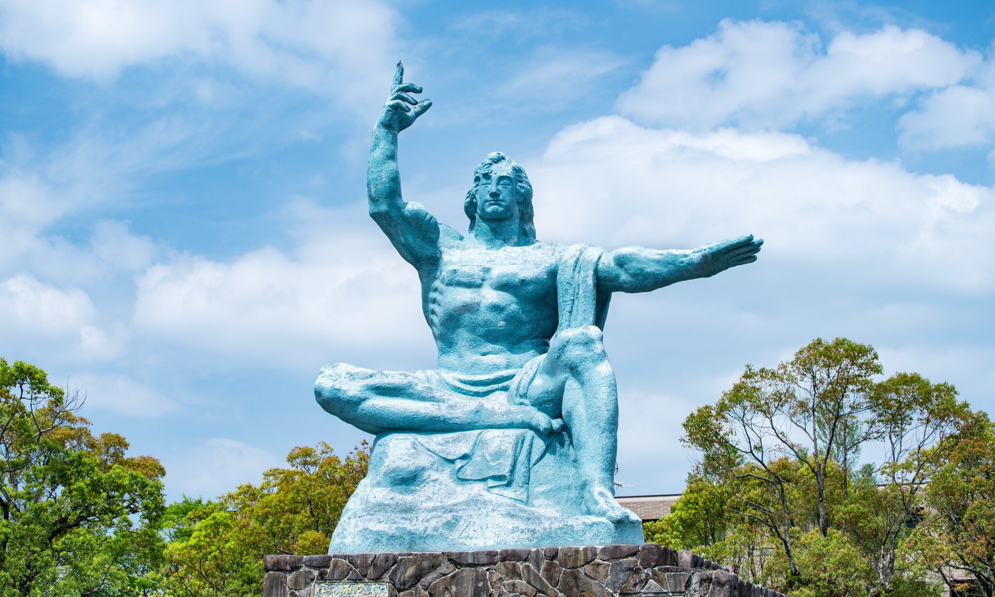 A large blue-green seated statue with one arm raised and one outstretched.