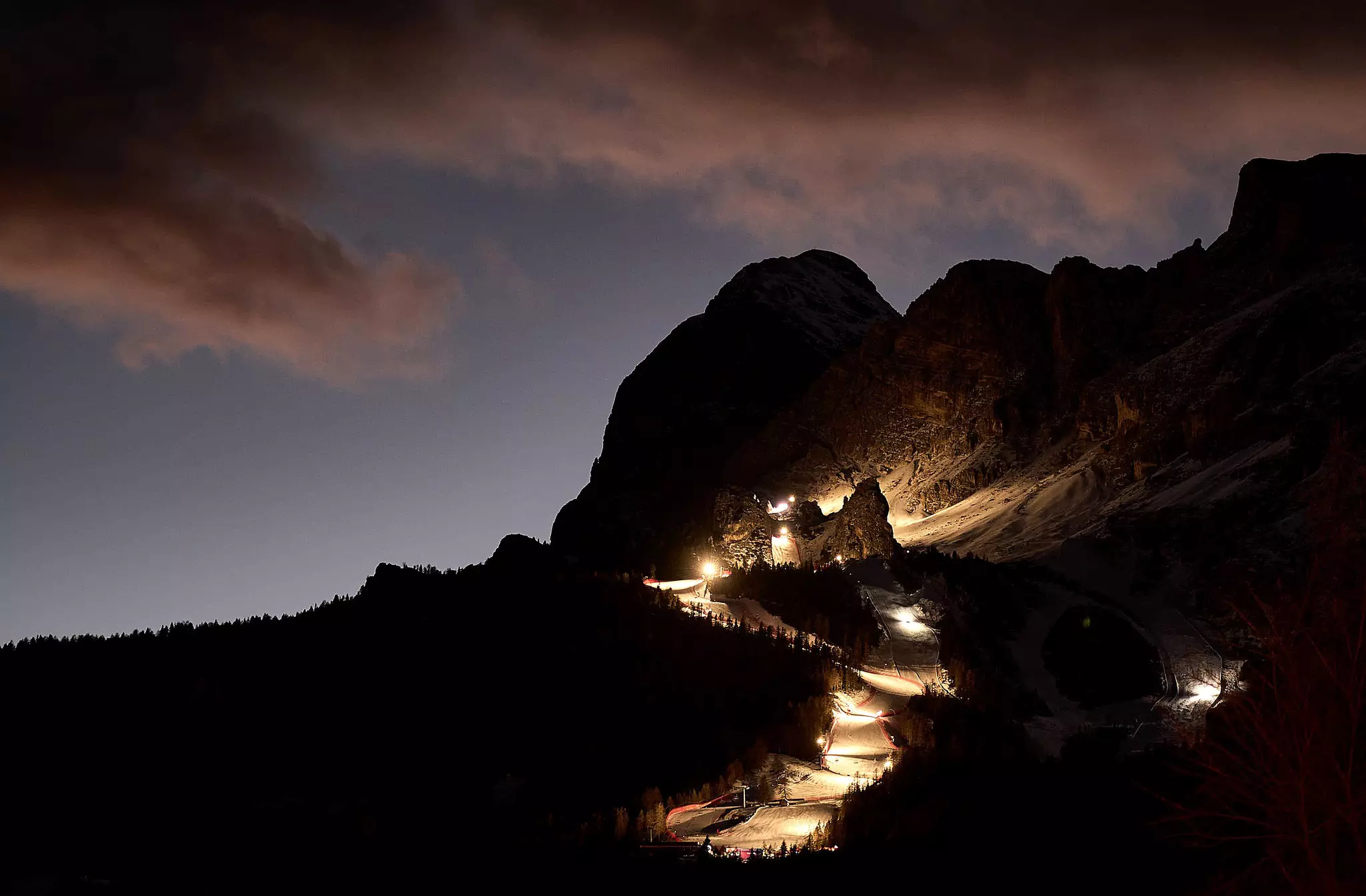 A view of a downhill ski run illuminated by floodlights at night. Mountains are seen in shadowy silhouette.