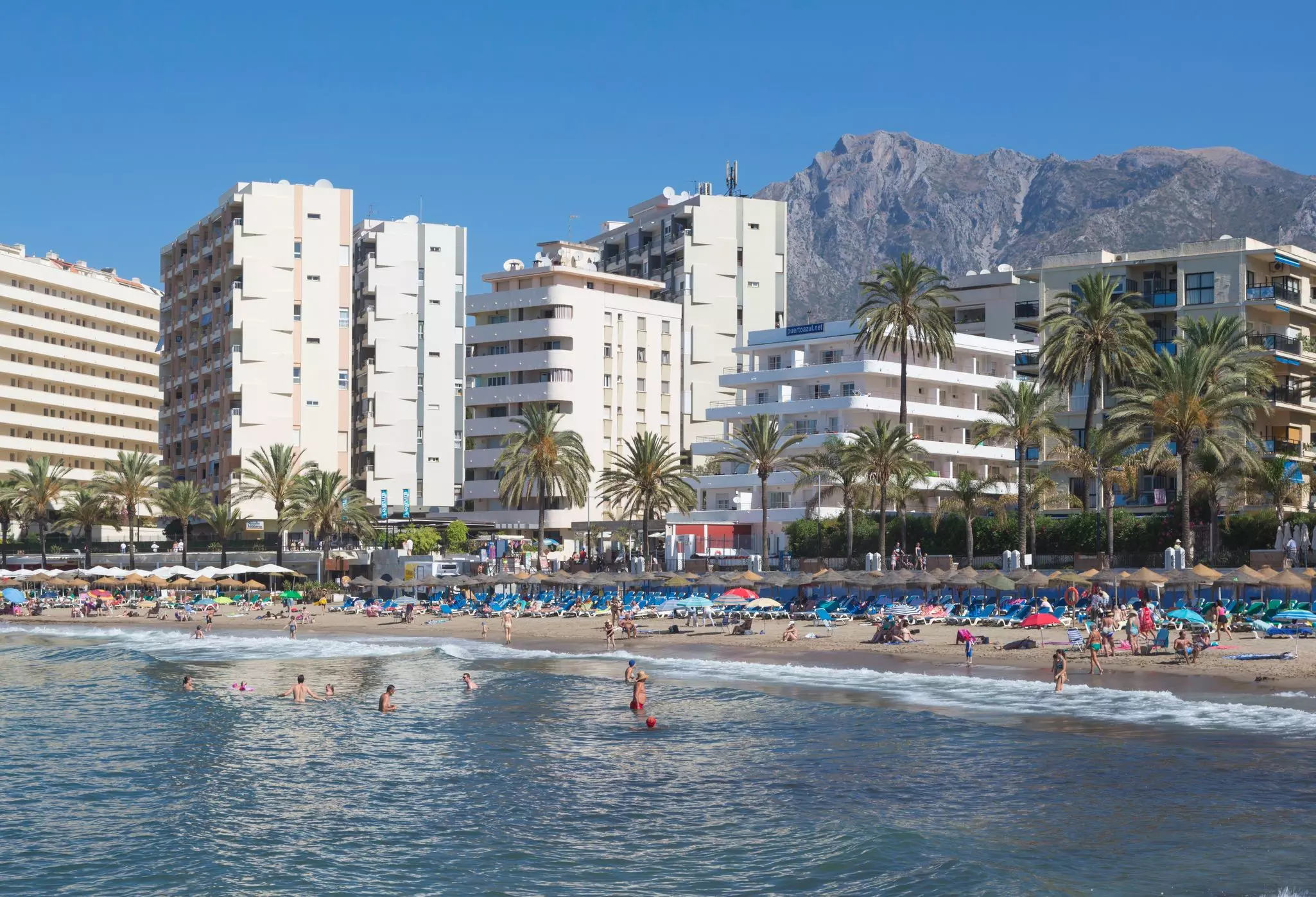 People on a beach with blue lounge chairs and in blue water in Spain. Palm trees separate the beach from white midrise buildings behind it; a mountain is in the background.