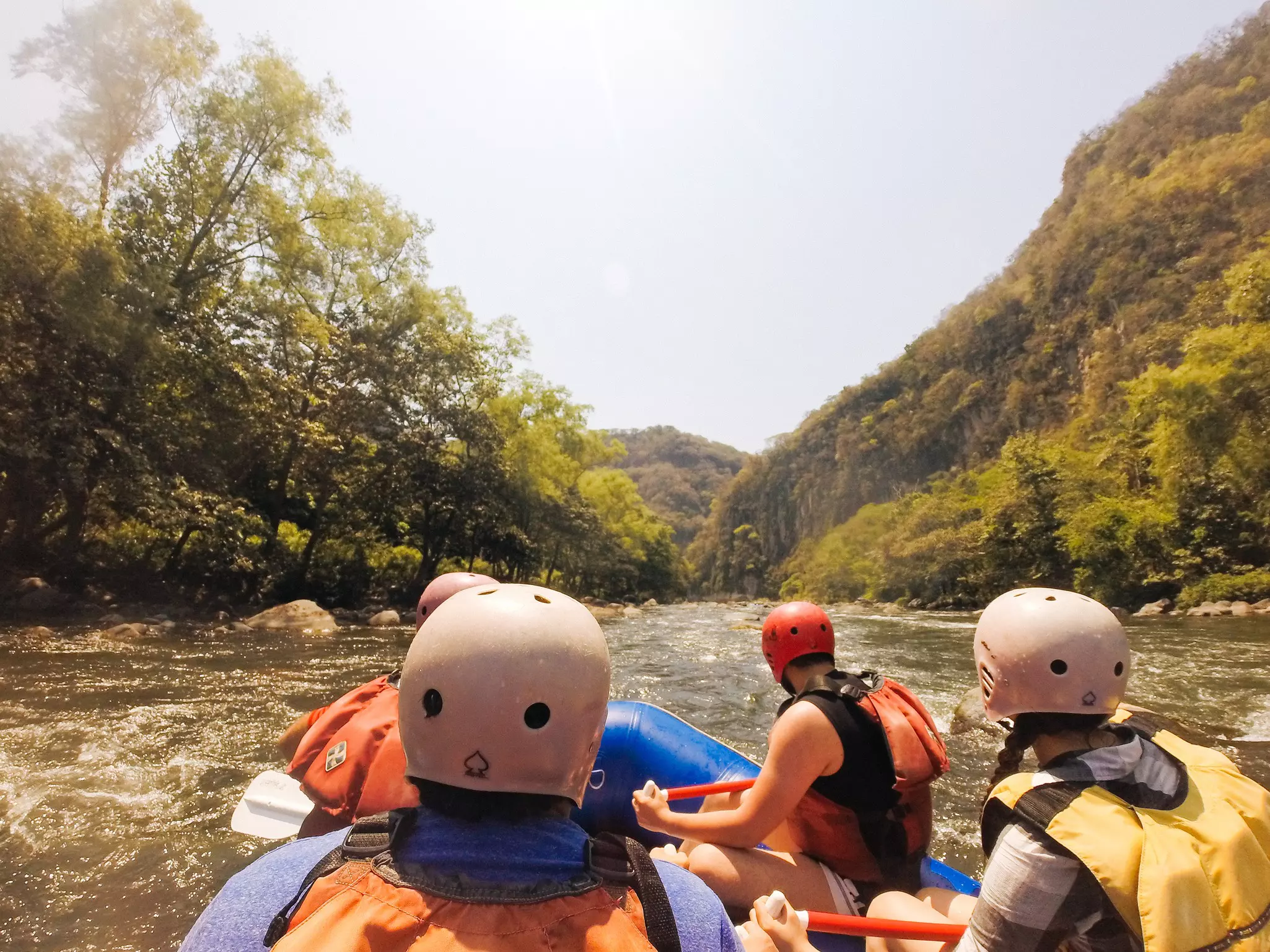 A view from behind of people in a raft on the white water of a river. Green hills rise from the banks of the river.