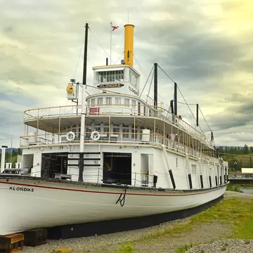 White SS Klondike sternwheeler with a yellow funnel resting on blocks beside the Yukon River in Whitehorse, Canada