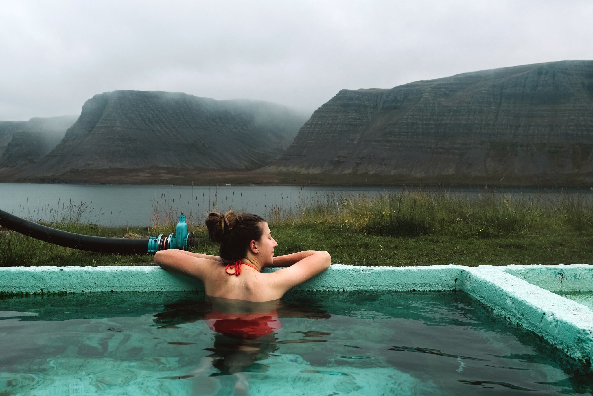 Woman in red swimsuit swimming in hot spring pool with view of morning foggy fjords and sea on Westfjords in Iceland