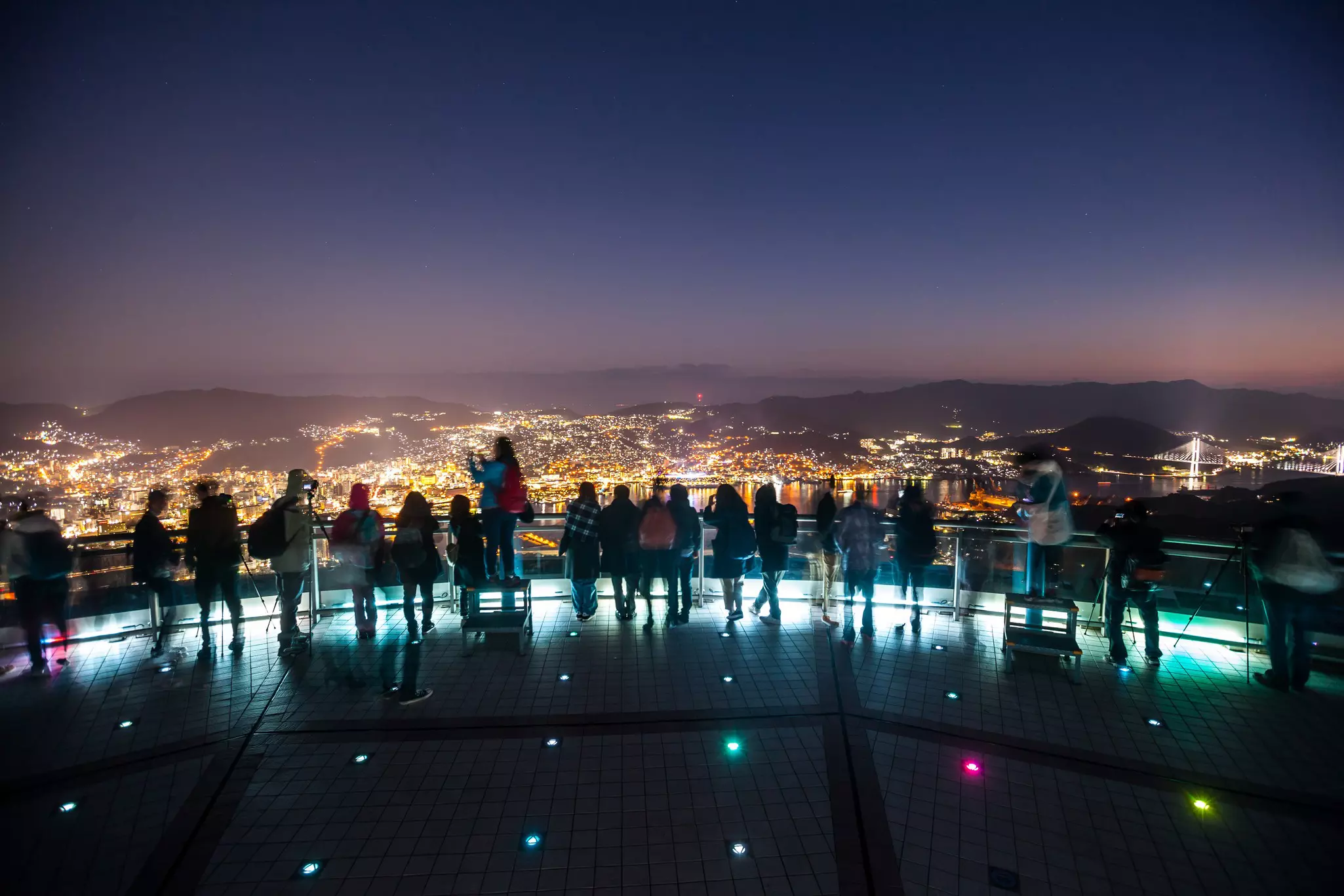 Tourists are silhouetted at night at an overlook, with the lights of Nagasaki below