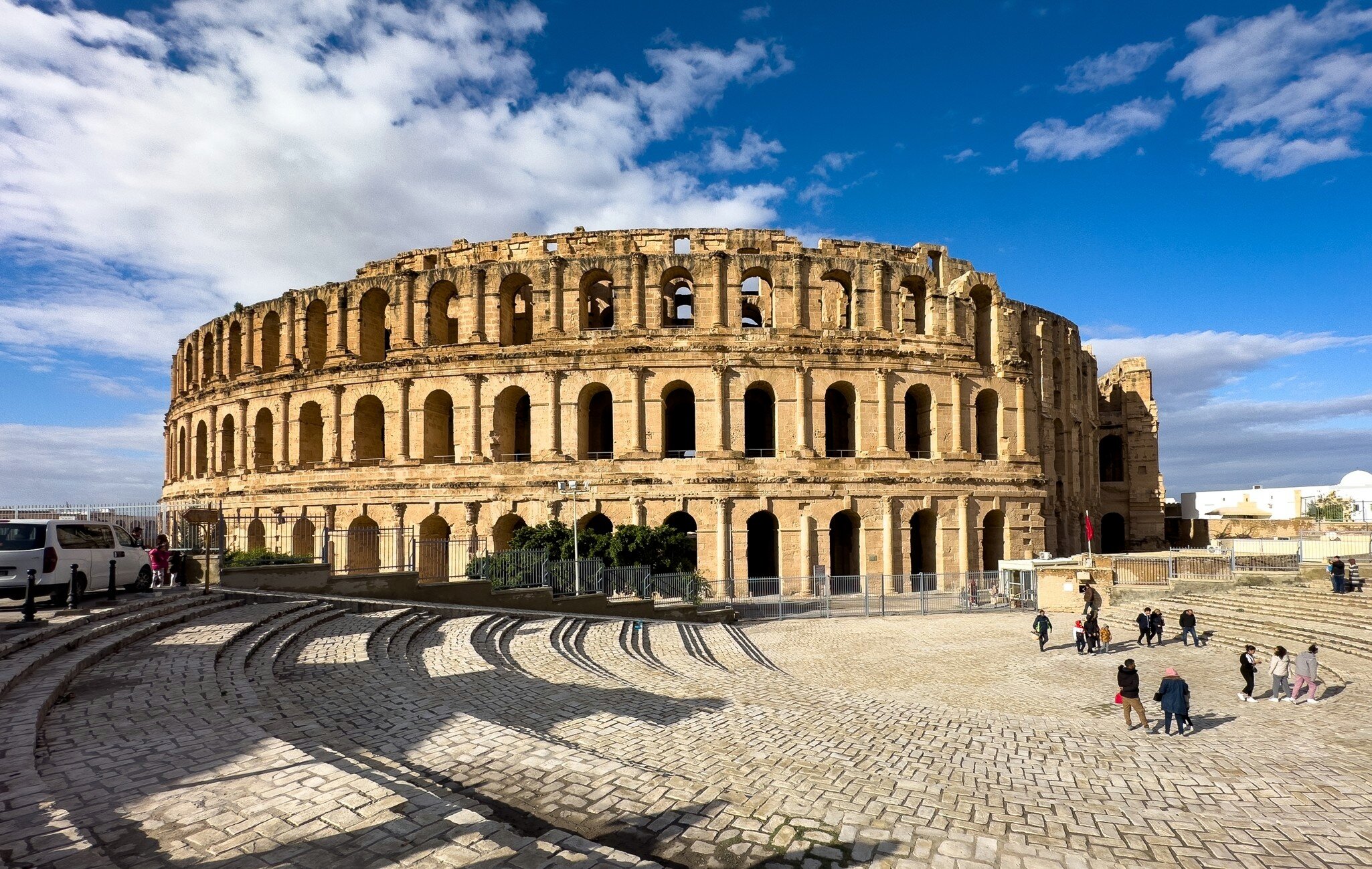 Tourists outside a well-maintained circular stone ruined amphitheater in a cobblestone square on a sunny day.