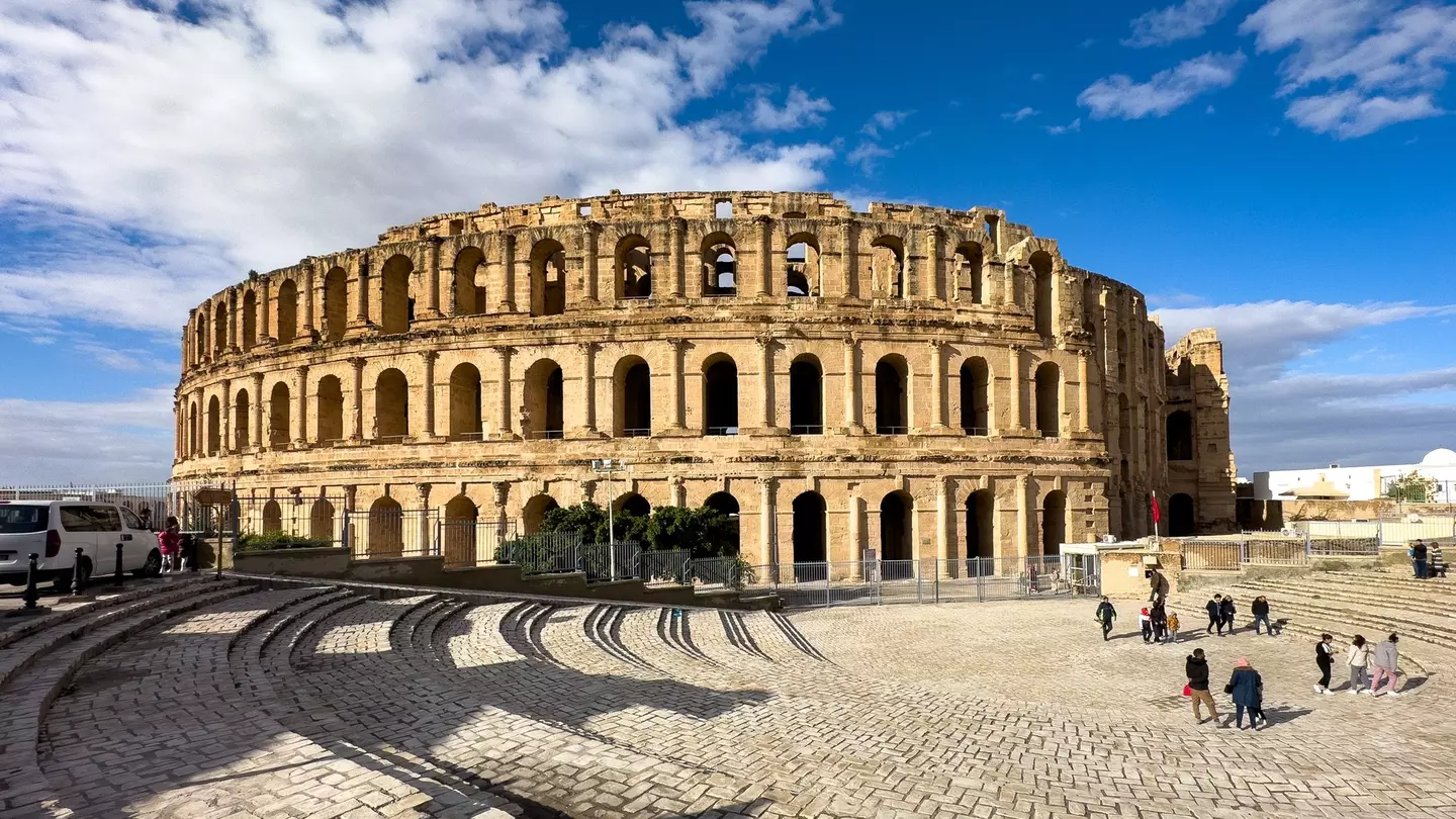 Tourists outside a well-maintained circular stone ruined amphitheater in a cobblestone square on a sunny day.