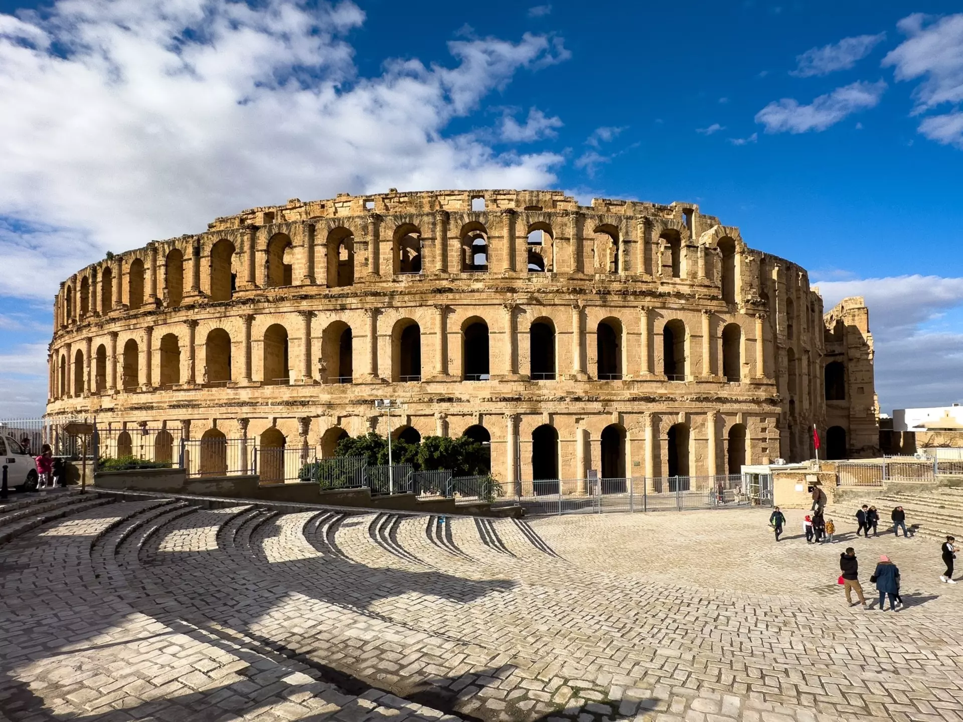 Tourists outside a well-maintained circular stone ruined amphitheater in a cobblestone square on a sunny day.