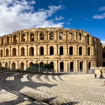 Tourists outside a well-maintained circular stone ruined amphitheater in a cobblestone square on a sunny day.