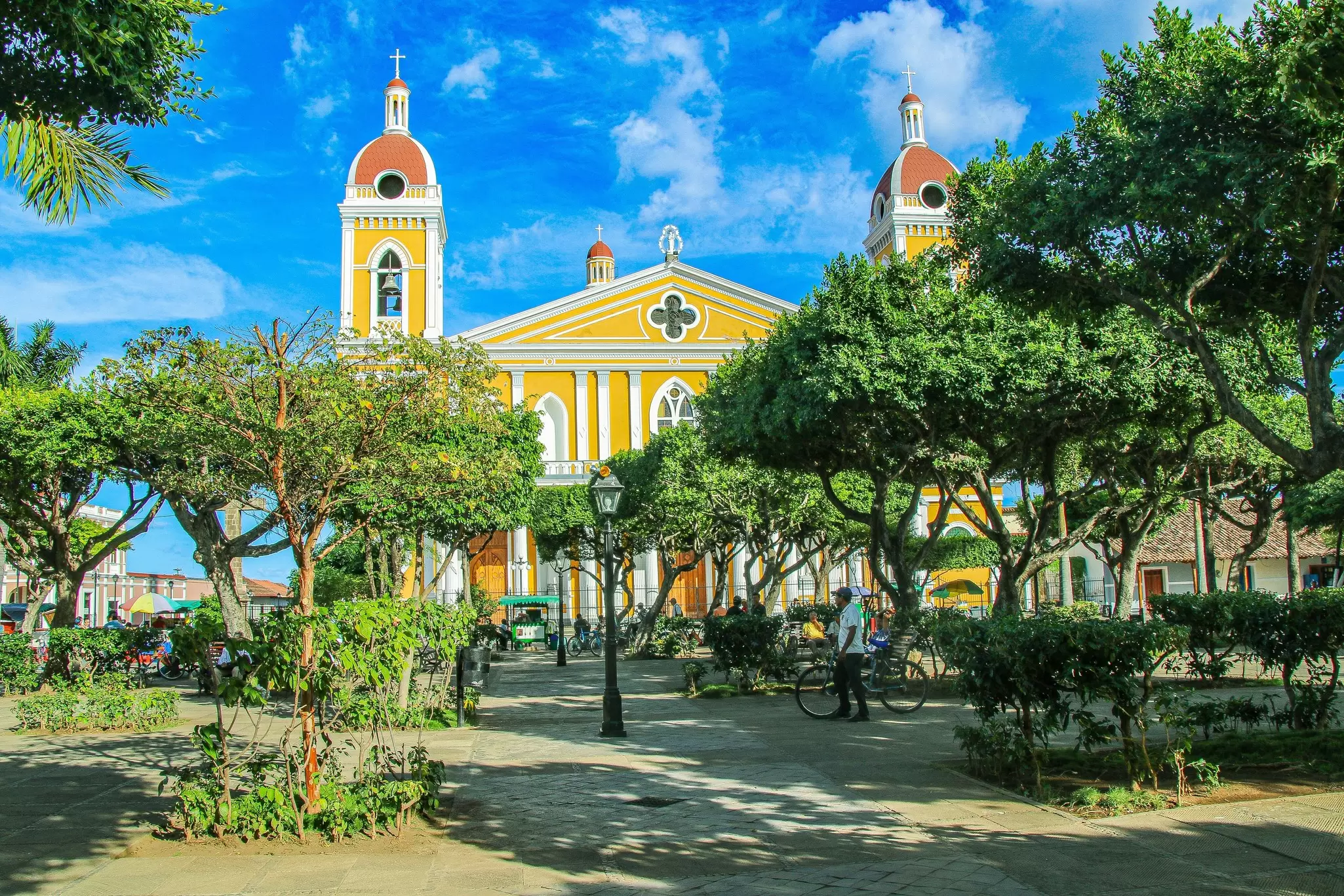 The wide facade of a church is seen from a square in front, with green trees shading the square, Granada, Nicaragua
