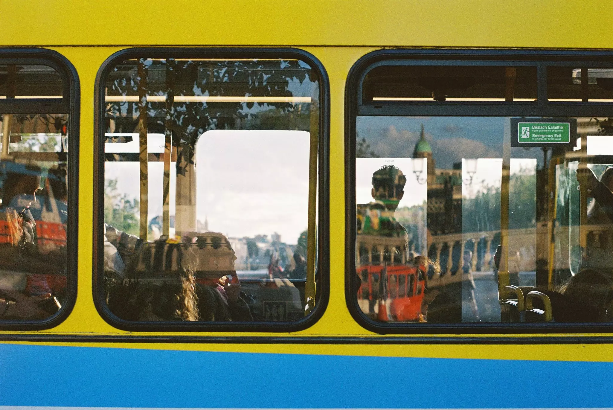 The side of the bus with three windows framing passengers with a window reflection of the city.
