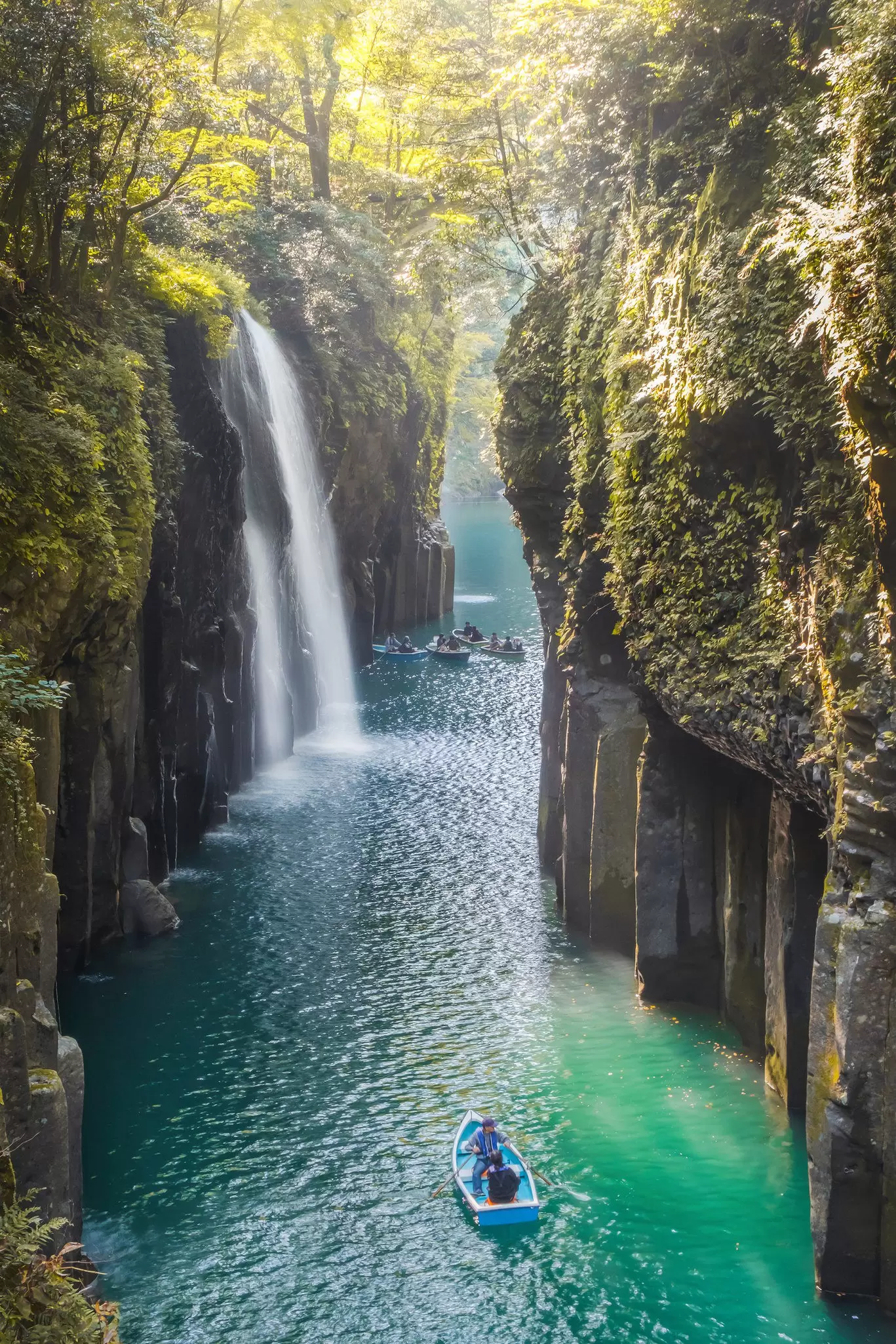 People in row boats sail through a narrow gorge with steep cliffs and a waterfall.