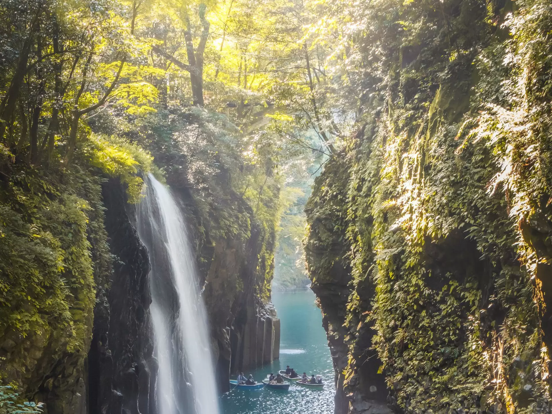People in row boats sail through a narrow gorge with steep cliffs and a waterfall.