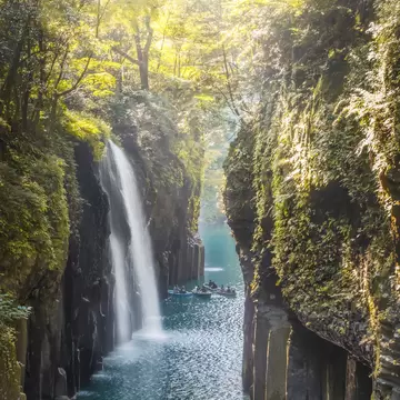 People in row boats sail through a narrow gorge with steep cliffs and a waterfall.