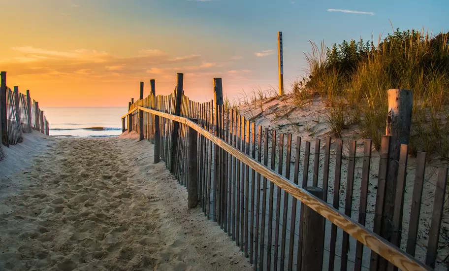 Weathered wooden fence lining a sandy path over dunes toward the ocean at sunset, marked by a Marine St sign