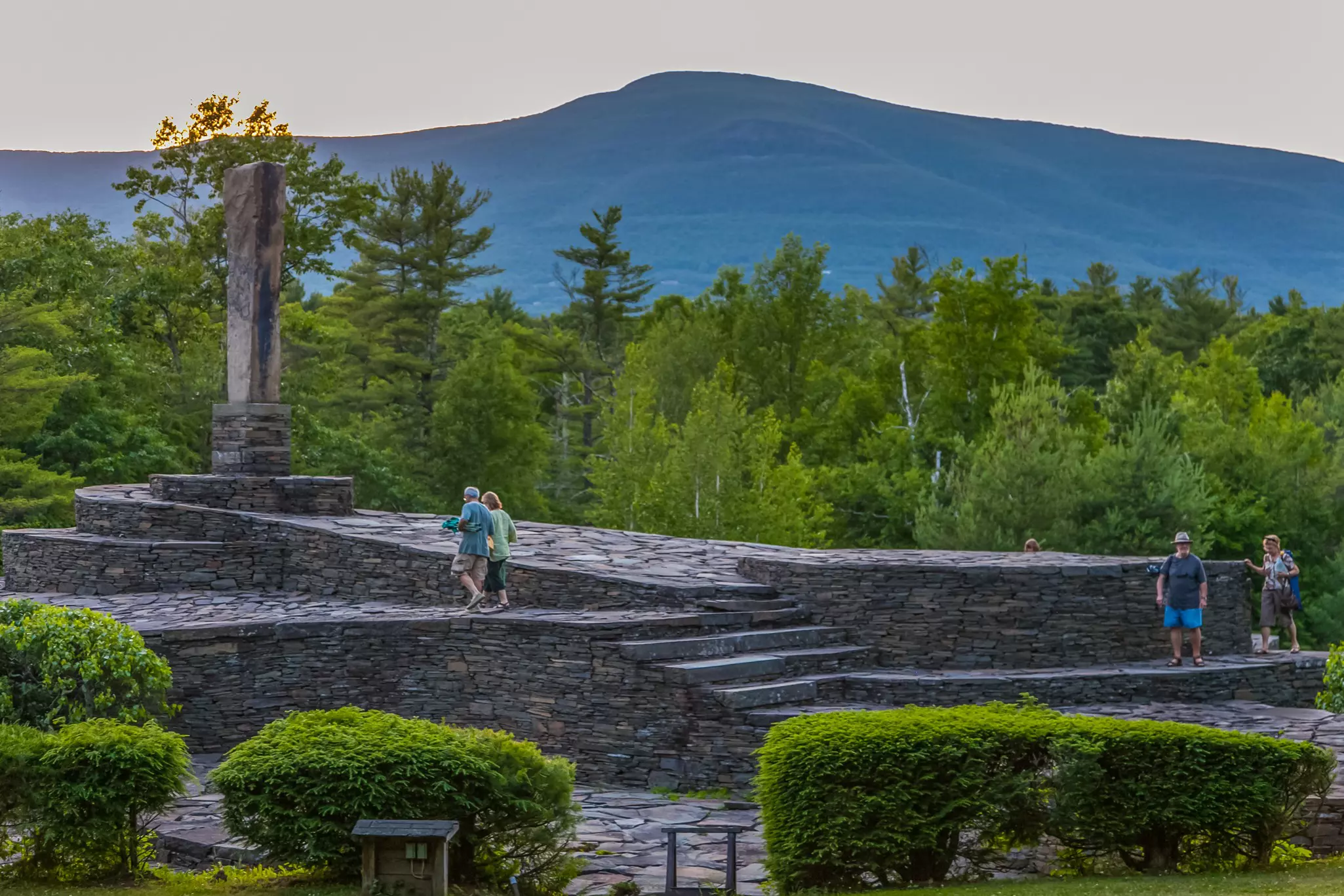 People walk on flagstones, part of an immersive outdoor artwork, surrounded by trees. A mountain is visible in the distance.