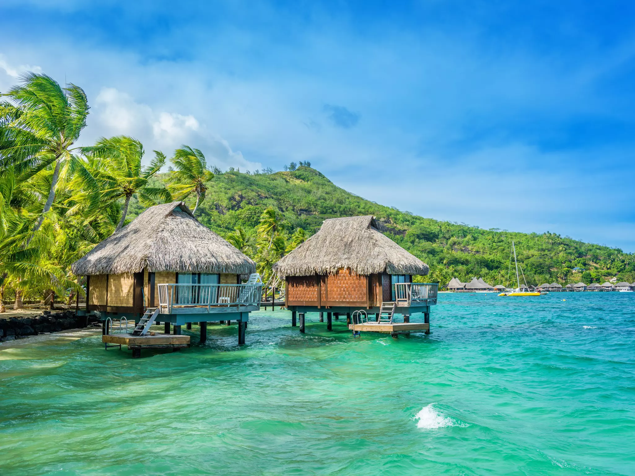 Two wooden thatched cabins on stilts stand in a turquoise lagoon on the edge of a lush island