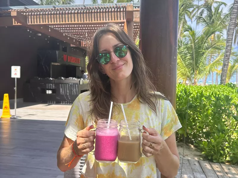 A person wearing sunglasses and a yellow tie-die shirt holds two glass mugs, one with pink contents and the other with brown contents.