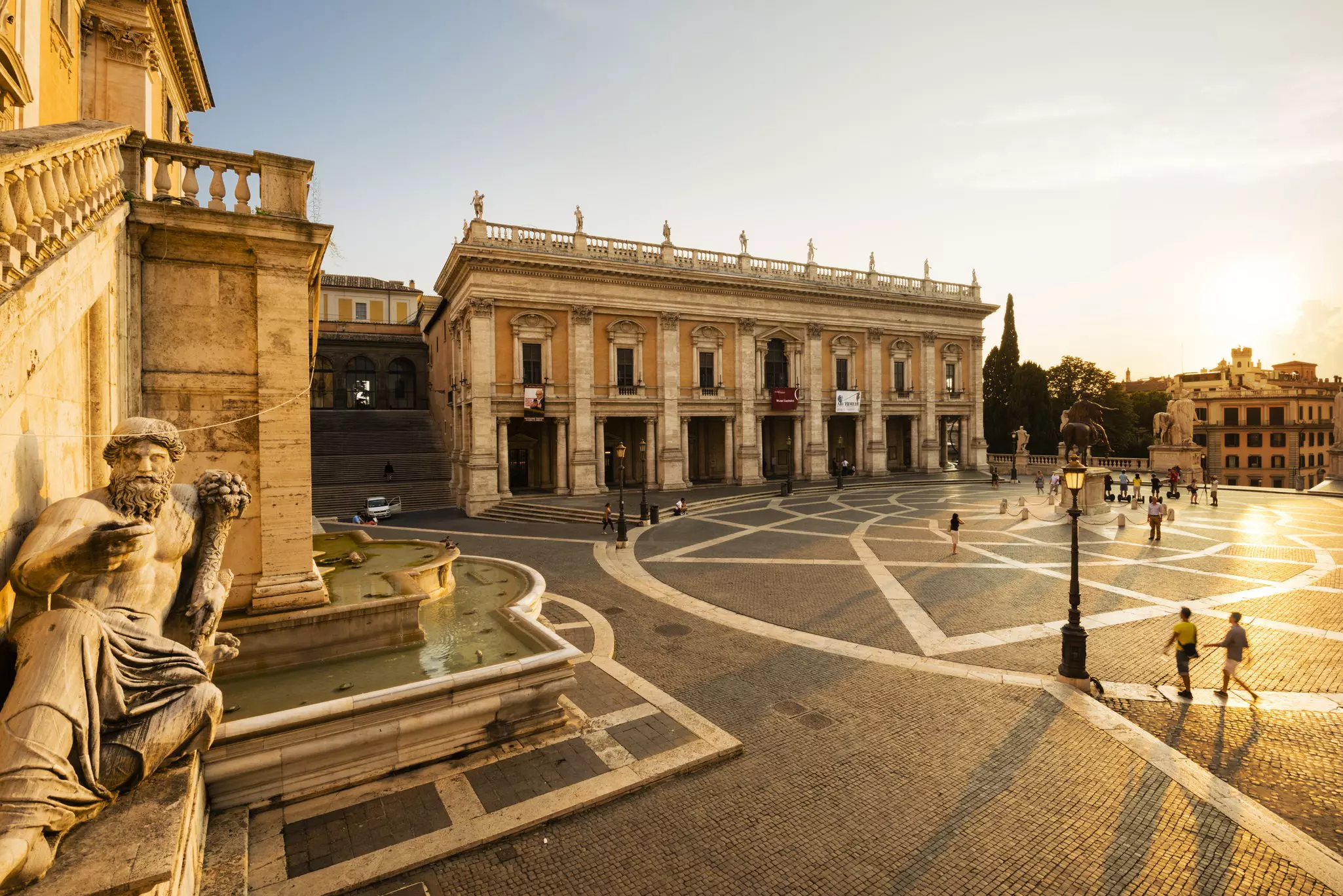 Evening sunlight illuminates the Piazza del Campidoglio with sculptures in the foreground and people walking through the square.