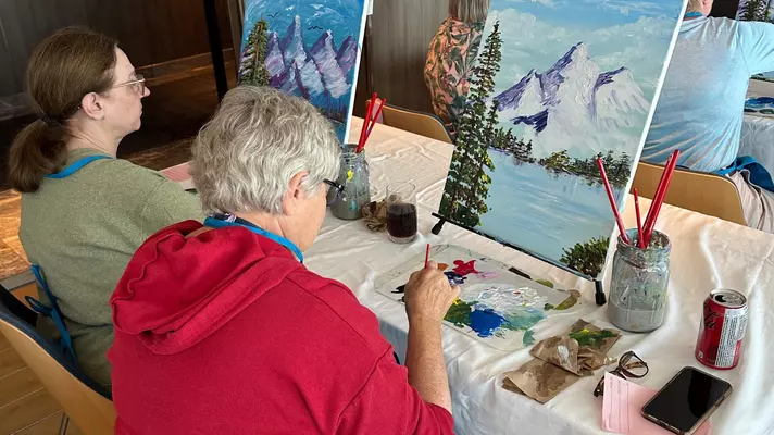 Two people sitting at a table painting on easels.