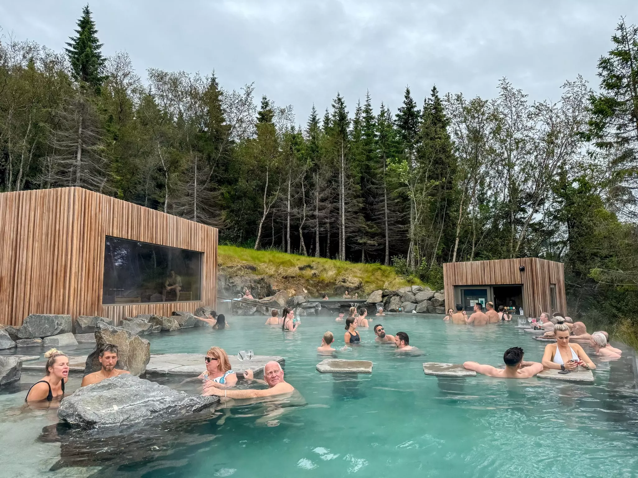 People soak in a spool at a hot springs resort. Wooden cabins are seen at the edge of a pool, with a pine forest beyond.