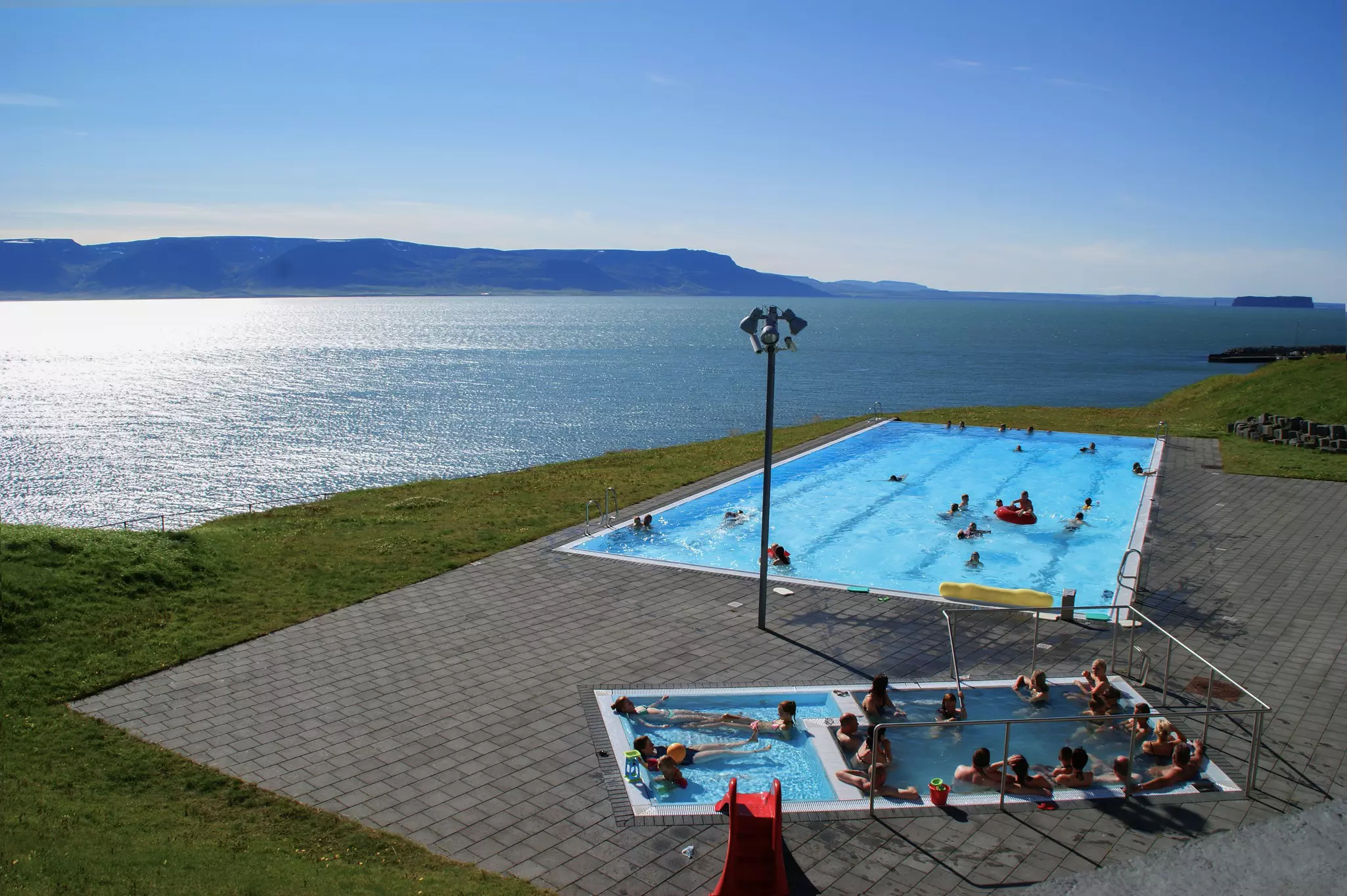 A large swimming pool and smaller soaking pools on a concrete patio alongside a fjord with mountains in the distance on a sunny day.