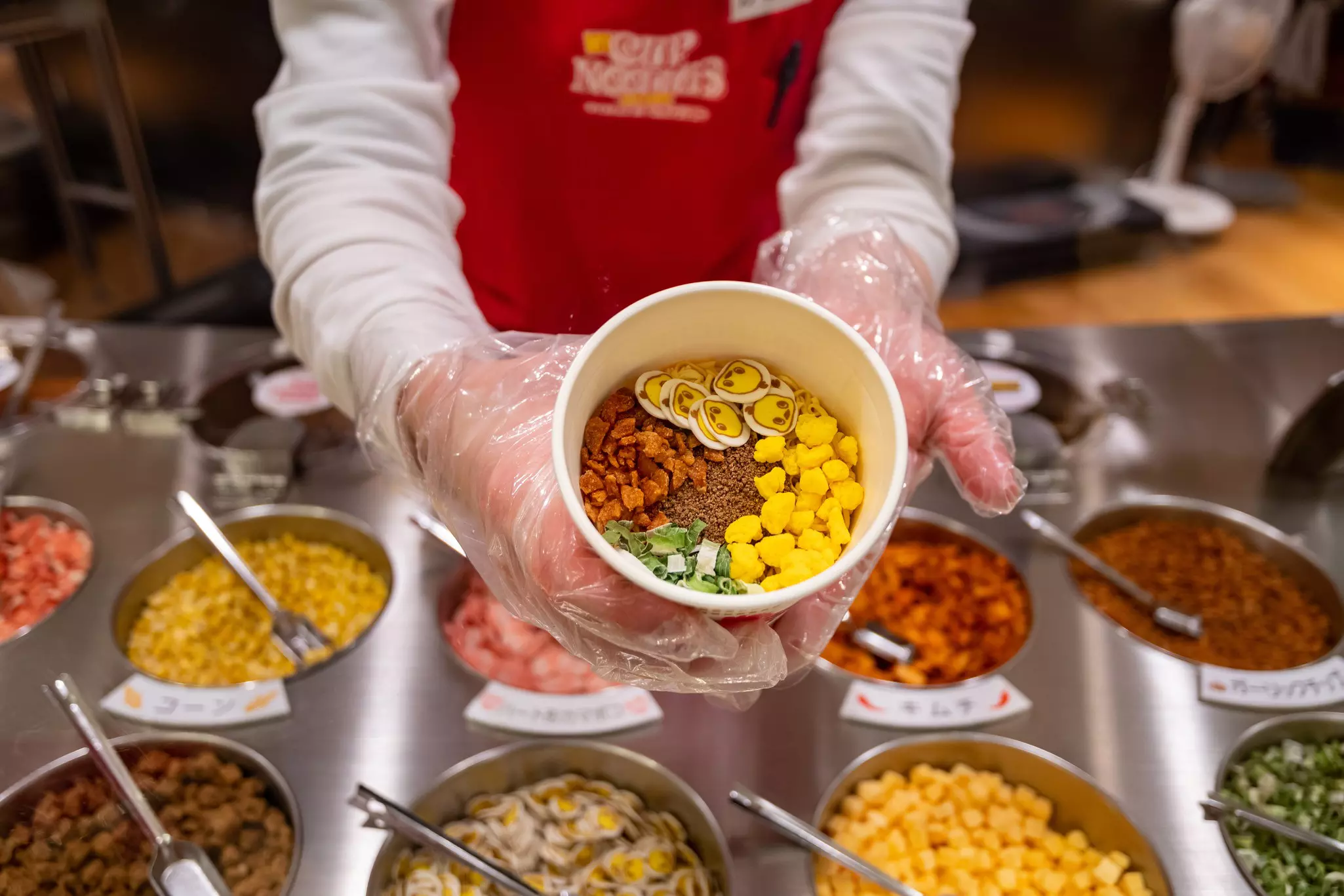 Woman's hands in clear plastic gloves holding an instant ramen container filled with dry ingredients.