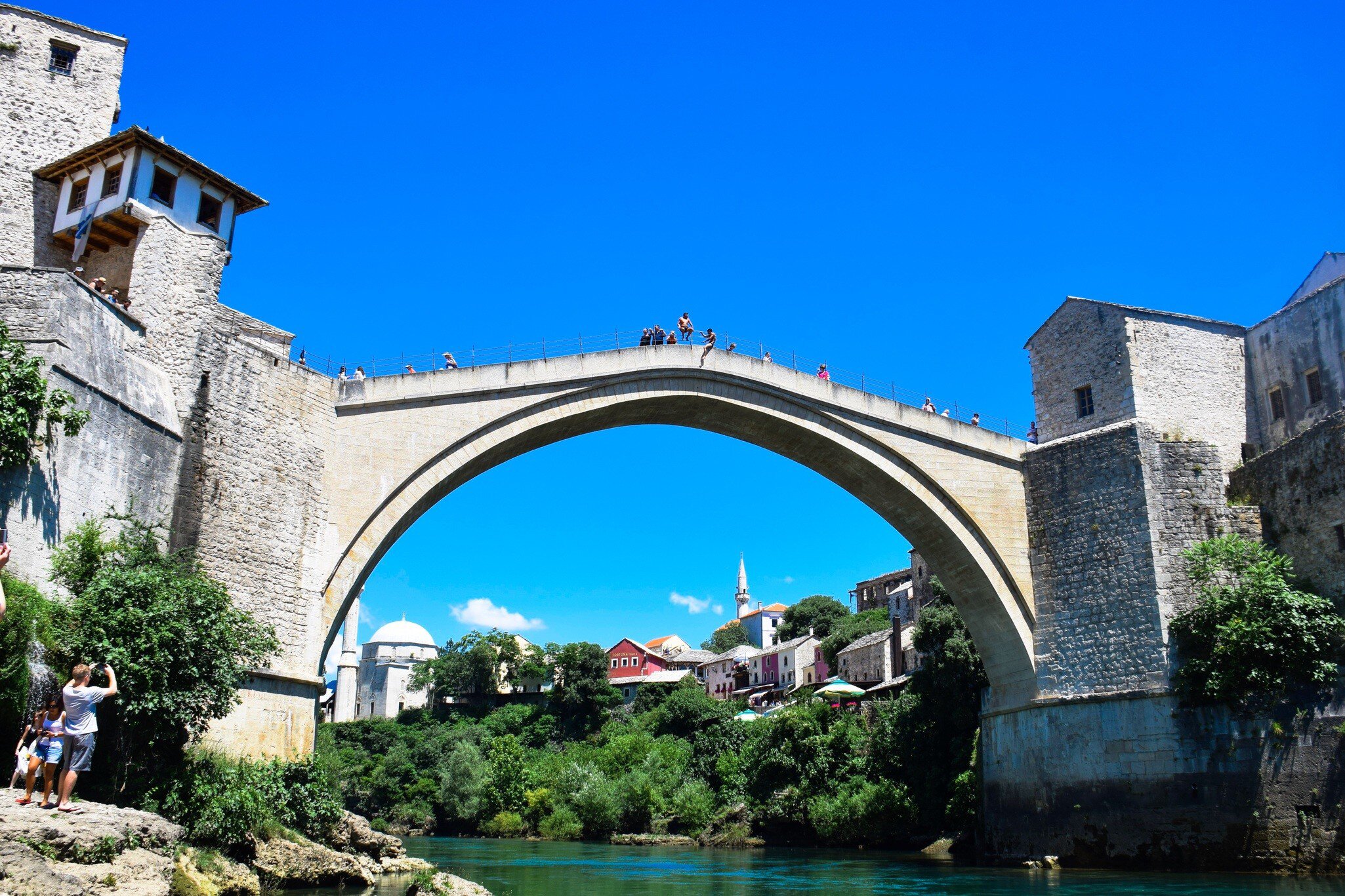 A single high stone arch spanning a green river between two fortified towers, onlookers along the parapet and a minaret in the old town beyond.